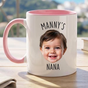 Puede incluir: Taza de cerámica blanca con interior y asa rosas. La taza presenta la cara sonriente de un bebé y el texto "Manny's Nana" en negro. Un libro y un paño doblado están en el fondo.