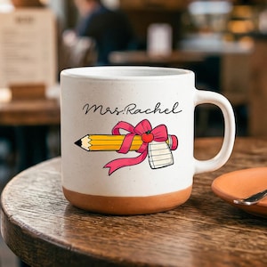 May include: A white ceramic mug with a speckled finish and a terracotta-colored base. The front features the text Mrs. Rachel in black script above a graphic of a yellow pencil tied with a pink ribbon and a small red apple, next to a notepad.