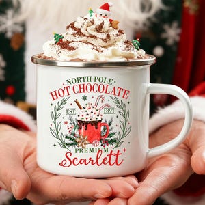 Könnte beinhalten: Ein weißer Emaillebecher gefüllt mit heißer Schokolade und Schlagsahne, garniert mit einer Santa-Figur und Streuseln. Auf dem Becher steht "North Pole Hot Chocolate Premium Scarlett" mit einem festlichen Design.