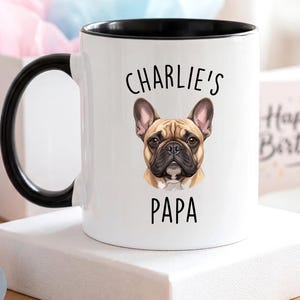 May include: White ceramic mug with a black handle and rim, featuring a French Bulldog illustration and the text "Charlie's Papa". A cupcake with blue frosting and a lit candle is in the background.