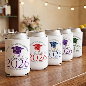 Puede incluir: Enfriadores de latas blancos con diseños de gorras de graduación y el texto "Class of 2026" y "Class of 2027". Cada enfriador tiene una gorra de graduación de diferente color y texto personalizado. Los enfriadores están sobre una superficie de madera.