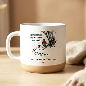 Può includere: Tazza in ceramica bianca con base beige, con un'illustrazione stravagante di due figure che camminano su un sentiero. La tazza presenta la scritta "NEVER FORGET THE DIFFERENCE YOU MAKE" e "mrs. smith" con un cuore rosso.