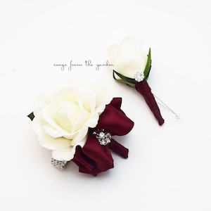 Puede incluir: Dos boutonnières de rosa blanca con cinta burdeos y detalles de pedrería. La boutonnière de la izquierda tiene un lazo y la boutonnière de la derecha es una sola rosa.