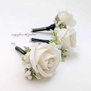 May include: Three white rose boutonnieres with green foliage and white baby's breath. The boutonnieres are attached to a dark green ribbon.
