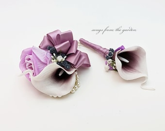 May include: A set of two purple and white flower boutonnieres with a satin bow and pearl accents. The boutonnieres are made with calla lilies and roses. "songs from the garden" is written in the top right corner of the image.