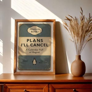 May include: A framed print with the text "PLANS I'LL CANCEL" in a vintage book cover design. The print features a penguin illustration and the words "A Calendar Full of Regret." The frame is made of wood, and the print is displayed on a wooden cabinet.