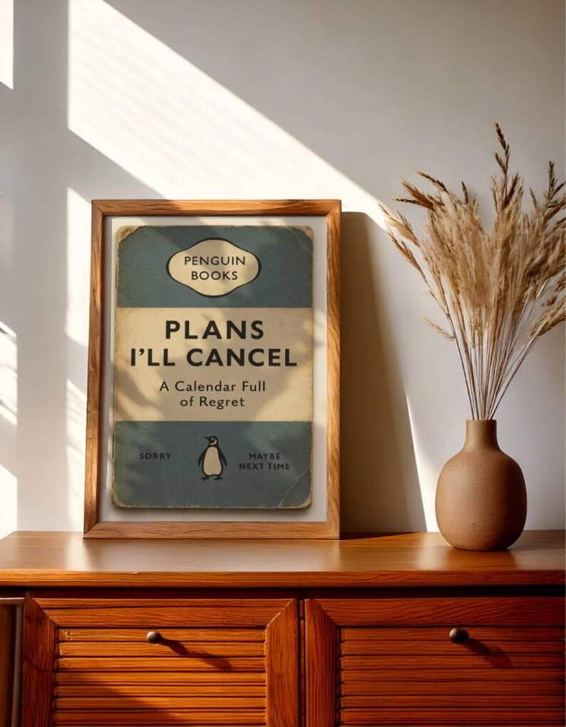 May include: A framed print with the text "PLANS I'LL CANCEL" in a vintage book cover design. The print features a penguin illustration and the words "A Calendar Full of Regret." The frame is made of wood, and the print is displayed on a wooden cabinet.