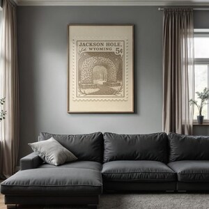 May include: A framed vintage-style print featuring a sepia-toned illustration of a tunnel with the text "Jackson Hole, Wyoming 5&cent;". The artwork hangs above a dark gray sectional sofa with light gray throw pillows in a living room setting.
