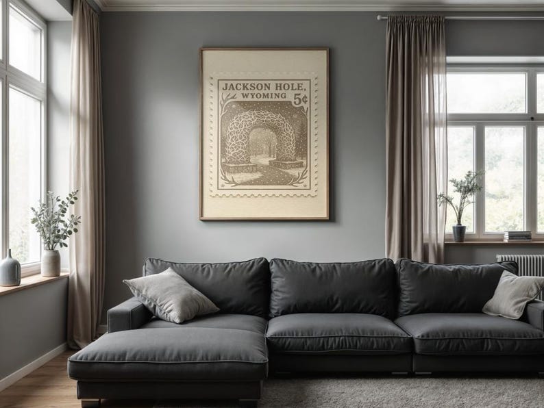 May include: A framed vintage-style print featuring a sepia-toned illustration of a tunnel with the text "Jackson Hole, Wyoming 5&cent;". The artwork hangs above a dark gray sectional sofa with light gray throw pillows in a living room setting.