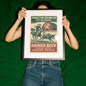 May include: Framed vintage-style poster featuring a rodeo scene with a cowboy on a horse and a bull. The poster has text that reads "Pendleton Round-Up" and "Rainier Beer - Since 1878 - Oregon Proud." The poster is held by a person wearing blue jeans.