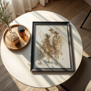 May include: A framed artwork with a watercolour painting of three figures wearing hats. The artwork is in a black frame and rests on a white round table. A gold tray with a small vase and a dark bottle sits on the table.