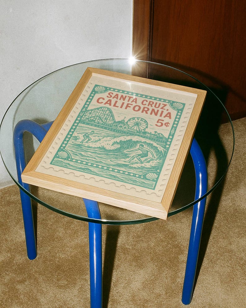 Może przedstawiać: Oprawiony wydruk z motywem vintage z Santa Cruz w Kalifornii, przedstawiający surfera i elementy parku rozrywki. Wydruk jest w jasnej drewnianej ramie i spoczywa na szklanym stole z niebieskimi nogami.