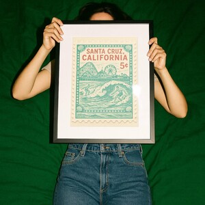 Może przedstawiać: Oprawiony wydruk z projektem znaczka pocztowego w stylu vintage. Znaczek głosi "Santa Cruz, California" i przedstawia surfer&oacute;w, kolejkę g&oacute;rską i diabelski młyn. Wydruk jest trzymany na zielonym tle.