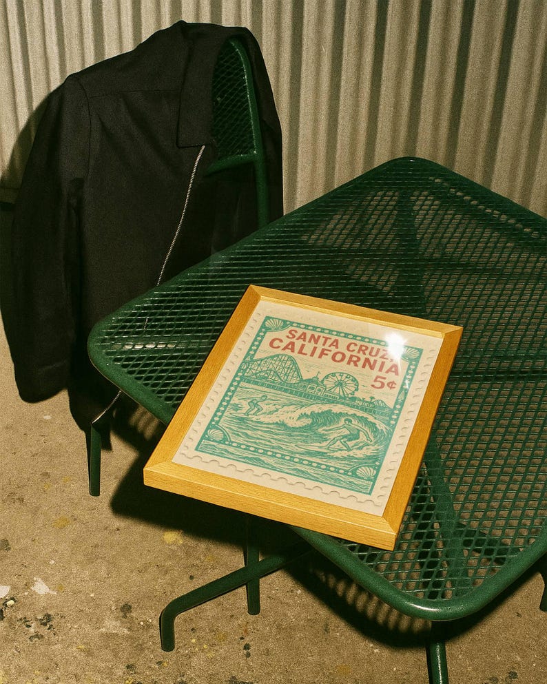 Może przedstawiać: Oprawiony wydruk z motywem vintage Santa Cruz, Kalifornia, przedstawiający kolejkę g&oacute;rską, diabelski młyn i surfer&oacute;w. Wydruk leży na zielonym metalowym stoliku. Czarna kurtka wisi na zielonym krześle.