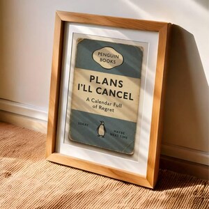 May include: A framed print with a light wood frame. The print features a vintage-style book cover design with the text "PLANS I'LL CANCEL" and a penguin illustration. The background is a light blue and beige color scheme.