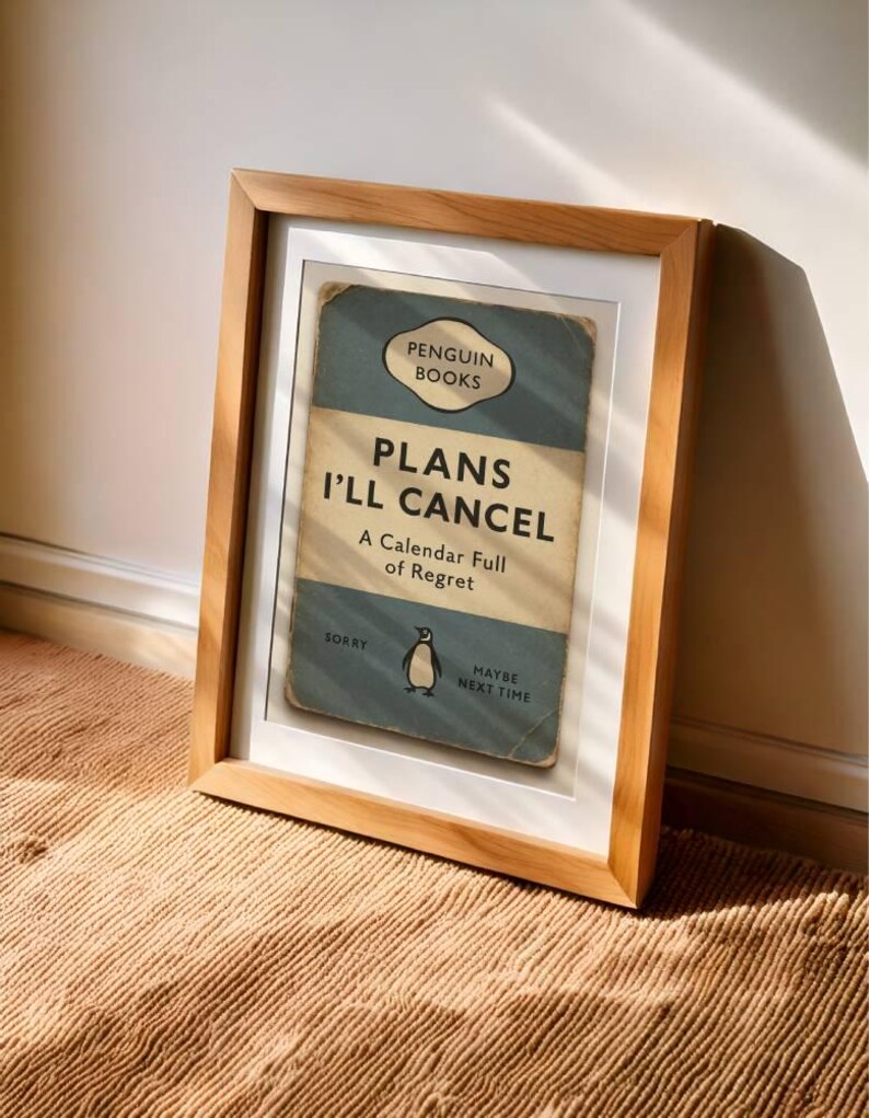 May include: A framed print with a light wood frame. The print features a vintage-style book cover design with the text "PLANS I'LL CANCEL" and a penguin illustration. The background is a light blue and beige color scheme.