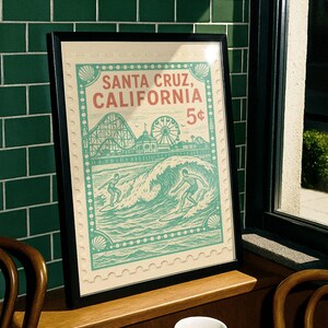 Może przedstawiać: Oprawiony wydruk z motywem vintage Santa Cruz, Kalifornia, z kolejką g&oacute;rską, diabelskim młynem i surferami. Grafika ma styl znaczka pocztowego z napisem "Santa Cruz, California 5&cent;". Grafika jest w czarnej ramie.