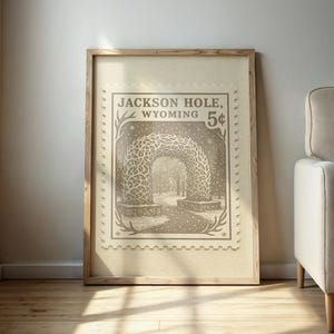 May include: A framed print featuring a vintage-style illustration of an archway in Jackson Hole, Wyoming, resembling a postage stamp. The artwork is in sepia tones and includes the text "Jackson Hole, Wyoming 5&cent;". The print is displayed against a neutral wall.