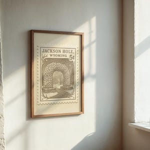 May include: Framed vintage-style print featuring a detailed illustration of an archway in Jackson Hole, Wyoming. The print has a sepia tone and is mounted in a wooden frame. The text "Jackson Hole, Wyoming 5&cent;" is visible.
