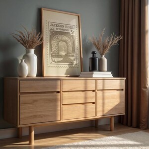 May include: A light-colored wooden sideboard with three drawers and two cabinet doors. The sideboard is topped with a framed print, vases, and books. The print reads "Jackson Hole Wyoming 5&cent;".