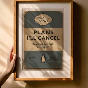 May include: Framed print with a vintage Penguin Books design. The print reads "PLANS I'LL CANCEL" with a penguin illustration. The frame is a light brown wood, and the print has a distressed, aged appearance.