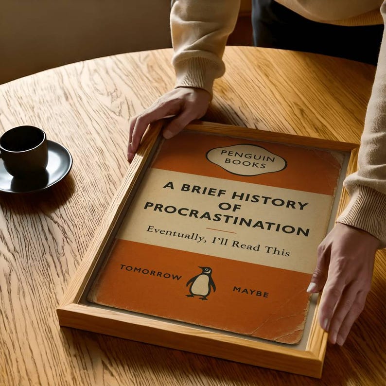 以下が含まれることがあります： ペンギンブックスの「A Brief History of Procrastination」というタイトルの額装された本の表紙。表紙はオレンジとクリーム色で、ペンギンのイラストが描かれています。フレームは明るい色の木材で作られています。