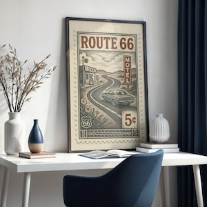 May include: A framed Route 66 art print. The vintage-style artwork features a winding road, a classic car, and the text "Route 66." The print is displayed on a white desk with decorative vases and a blue chair. The artwork is approximately 18 x 24 inches.