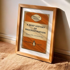 以下が含まれることがあります： ペンギンブックスの「A Brief History of Procrastination」というタイトルの本の表紙の額装プリント。表紙はオレンジとクリーム色で、ペンギンのイラストが描かれています。テキストは「Eventually, I'll Read This」と書かれています。