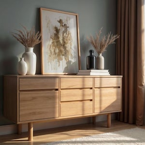 May include: A light oak sideboard with three drawers and three doors. The sideboard is topped with a framed abstract art print, vases, and books. The room has a neutral colour palette with a window and curtains.