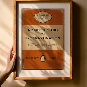 以下が含まれることがあります： 「A Brief History of Procrastination」というタイトルの本の表紙の額装されたプリント。Penguin Booksによる。表紙はオレンジとタンで、ペンギンのイラストと「Eventually, I'll Read This」というテキストが描かれています。フレームは木製です。