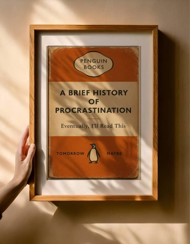 以下が含まれることがあります： 「A Brief History of Procrastination」というタイトルの本の表紙の額装されたプリント。Penguin Booksによる。表紙はオレンジとタンで、ペンギンのイラストと「Eventually, I'll Read This」というテキストが描かれています。フレームは木製です。