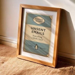 May include: A framed print of a book cover titled "Unsent Emails" by Penguin Books. The design features a vintage aesthetic with a light blue and beige color scheme. The frame is made of light-colored wood.