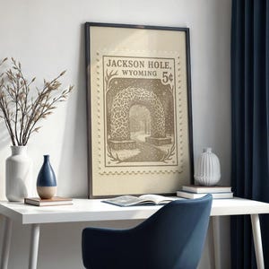 May include: Framed vintage-style print featuring a sepia-toned illustration of an archway in Jackson Hole, Wyoming, with the text "Jackson Hole, Wyoming 5&cent;". The print is displayed on a white desk with a blue chair, vases, and a book.