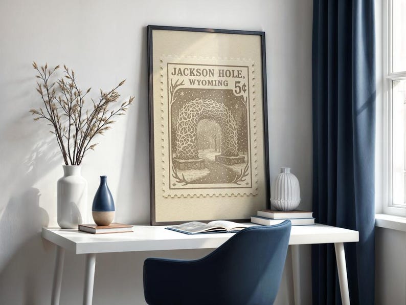 May include: Framed vintage-style print featuring a sepia-toned illustration of an archway in Jackson Hole, Wyoming, with the text "Jackson Hole, Wyoming 5&cent;". The print is displayed on a white desk with a blue chair, vases, and a book.