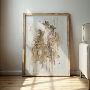May include: Abstract art print depicting three figures in cowboy hats, in shades of brown and beige. The artwork is framed in a light wood frame and leans against a neutral wall, creating a rustic aesthetic.