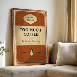 May include: A framed print featuring a vintage book cover design. The cover is orange and cream, with the text "TOO MUCH COFFEE" and "Anxiety in Every Sip." A penguin illustration is centered below the text. The frame is made of light-coloured wood.