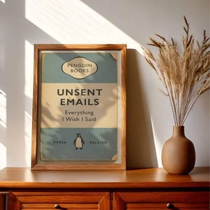 May include: A framed print with the text "UNSENT EMAILS Everything I Wish I Said" in a vintage book cover design. The print features a penguin illustration and the words "TYPED" and "DELETED". The frame is made of wood, and the print is displayed on a wooden cabinet.