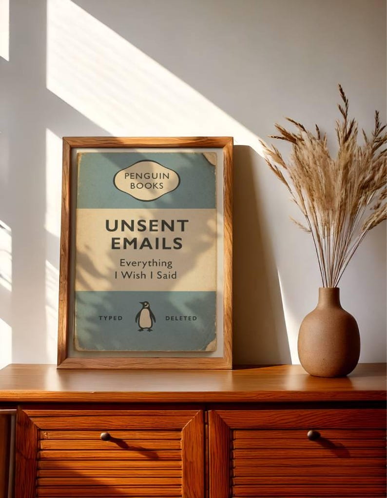 May include: A framed print with the text "UNSENT EMAILS Everything I Wish I Said" in a vintage book cover design. The print features a penguin illustration and the words "TYPED" and "DELETED". The frame is made of wood, and the print is displayed on a wooden cabinet.