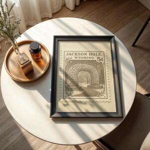 May include: A framed print featuring a vintage postage stamp design with the text "Jackson Hole, Wyoming 5&cent;". The print is in a black frame and rests on a white table. A gold tray with a small vase and a bottle is also on the table.