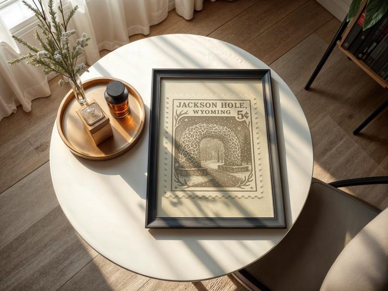 May include: A framed print featuring a vintage postage stamp design with the text "Jackson Hole, Wyoming 5&cent;". The print is in a black frame and rests on a white table. A gold tray with a small vase and a bottle is also on the table.