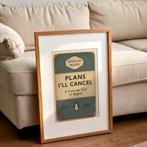 May include: A framed print featuring a vintage Penguin Books cover design. The cover reads "PLANS I'LL CANCEL" with the subtitle "A Calendar Full of Regret." The frame is light brown, and the print is set against a white mat.