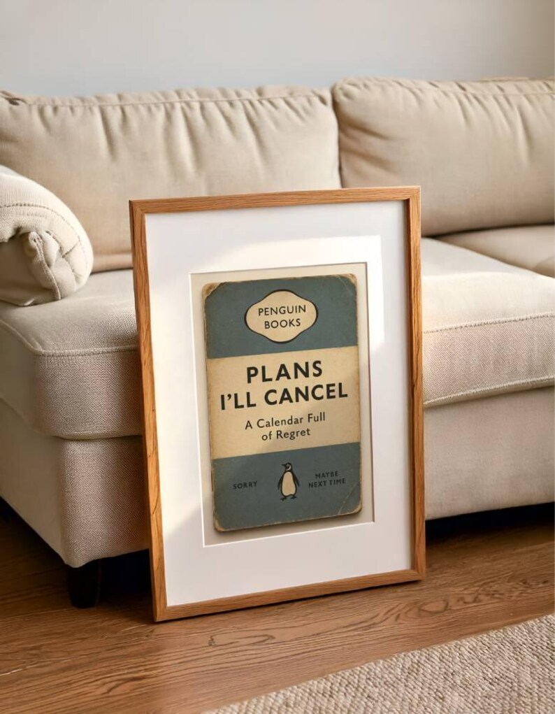 May include: A framed print featuring a vintage Penguin Books cover design. The cover reads "PLANS I'LL CANCEL" with the subtitle "A Calendar Full of Regret." The frame is light brown, and the print is set against a white mat.