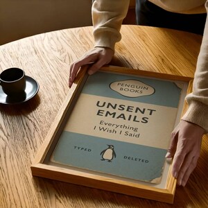 May include: A framed print with the text "Unsent Emails Everything I Wish I Said" from Penguin Books. The print has a light blue and beige background with a penguin logo. The frame is made of light brown wood.