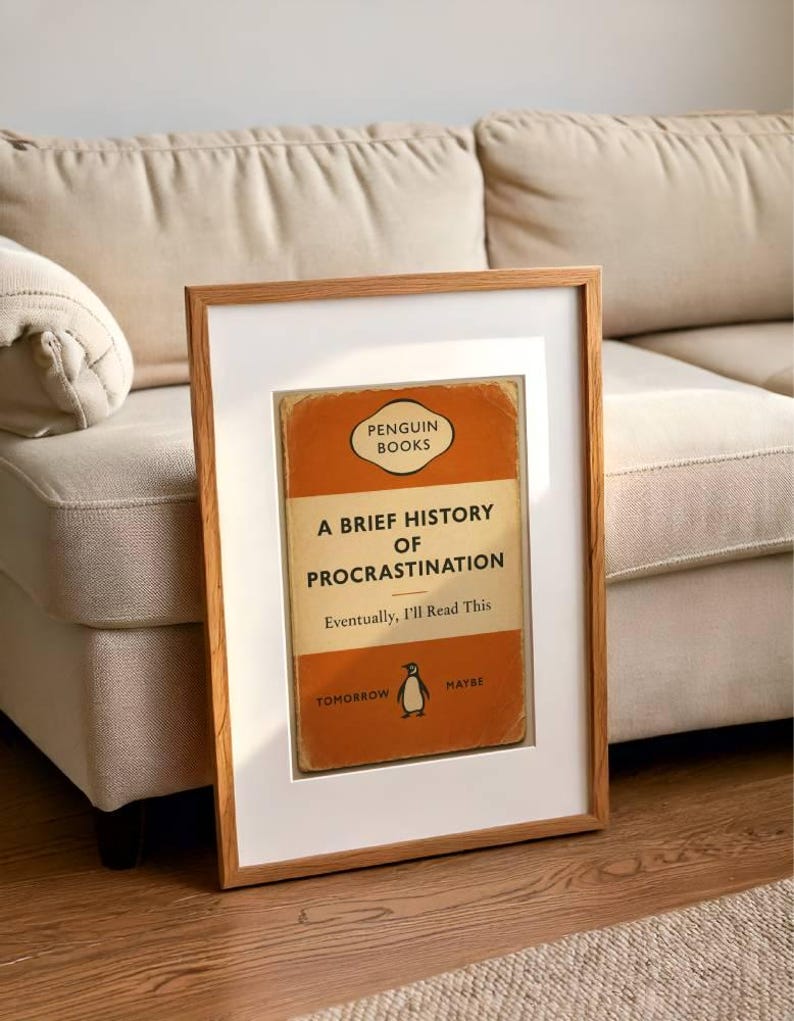 以下が含まれることがあります： 「A Brief History of Procrastination」というタイトルのペンギンブックスの書籍カバーの額装プリント。カバーはオレンジとクリーム色で、ペンギンのロゴが入っています。フレームはライトブラウンです。