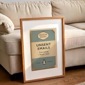 May include: Framed print featuring a vintage Penguin Books cover design. The cover is light blue and tan, with the title "Unsent Emails: Everything I Wish I Said." The frame is a light brown wood, and the print is matted in white.