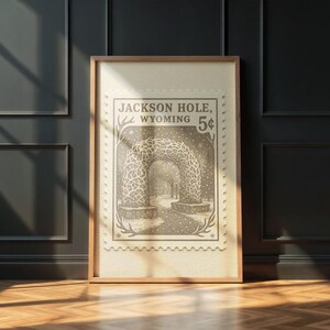 May include: Framed print of a vintage postage stamp design featuring an archway in Jackson Hole, Wyoming. The stamp is in sepia tones with the text "Jackson Hole, Wyoming 5&cent;". The print is in a wooden frame.