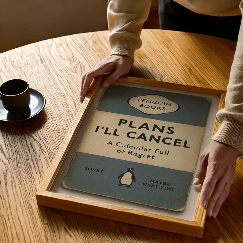 May include: A framed print of a Penguin Books book cover titled "Plans I'll Cancel: A Calendar Full of Regret." The cover is blue and white with a penguin illustration. The frame is light brown.