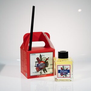 Puede incluir: Una caja de cartón roja con asa y una botella de vidrio transparente con tapa negra. La botella contiene un líquido amarillo y tiene una etiqueta con el texto "Pelara London Garden" y una imagen de monumentos de Londres. La caja tiene una etiqueta similar con el texto "Pelara" y una imagen de monumentos de Londres.
