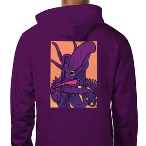Puede incluir: Sudadera con capucha morada con un estampado gráfico de una criatura alienígena morada con una lengua rosa en la espalda. El alienígena está en un fondo rosa y naranja.