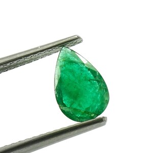 May include: A pear-shaped, faceted emerald gemstone held by metal tweezers. The gemstone is a vibrant green color and has visible internal inclusions. The tweezers are silver-colored and provide a contrasting background.